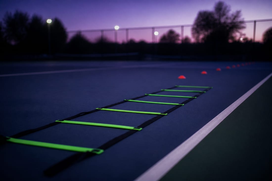 An agility ladder laid flat on a tennis court beside the baseline with cones spaced beyond it