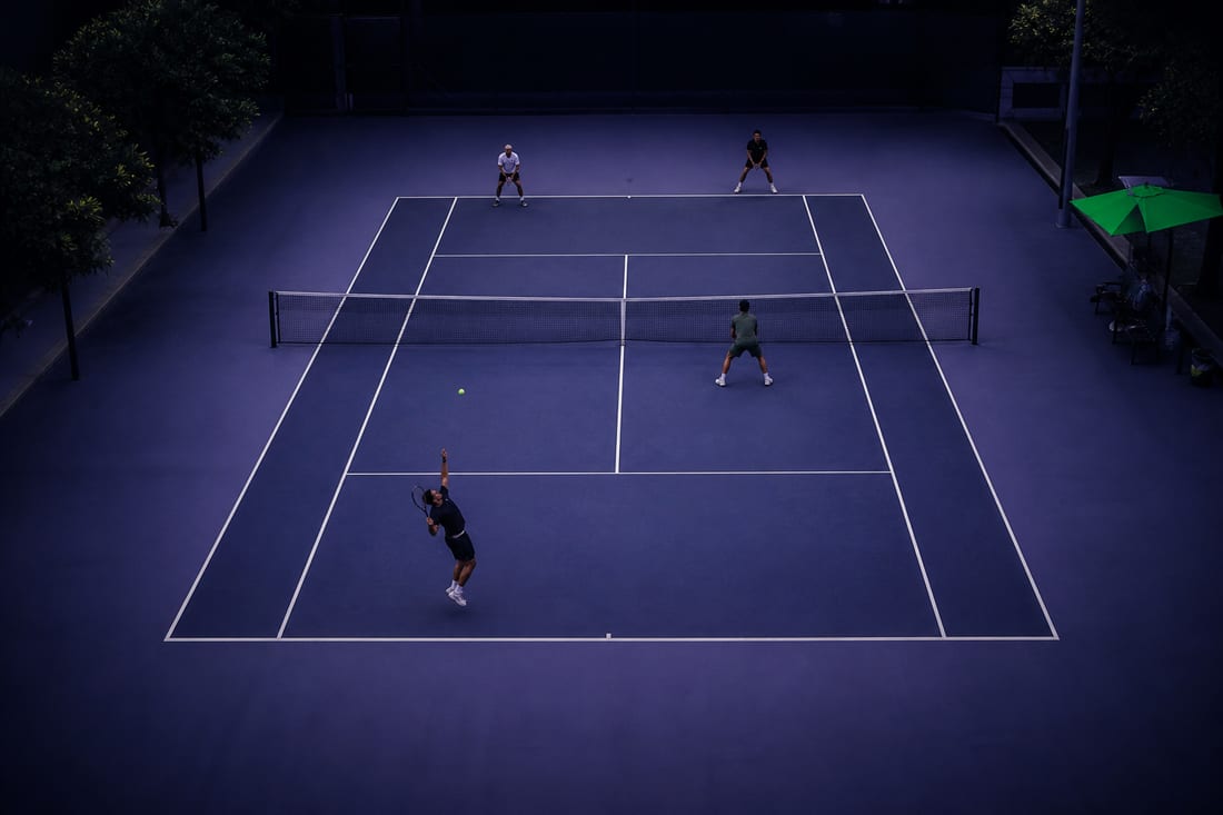 A doubles court from overhead with four distant players in the standard formation: server at the baseline, partner at the net, two receivers opposite