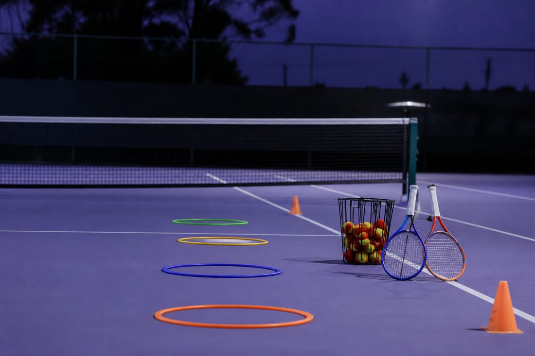 A junior coaching setup on court with throw-down hoops in a row, cones, short junior racquets and a basket of red low-compression balls