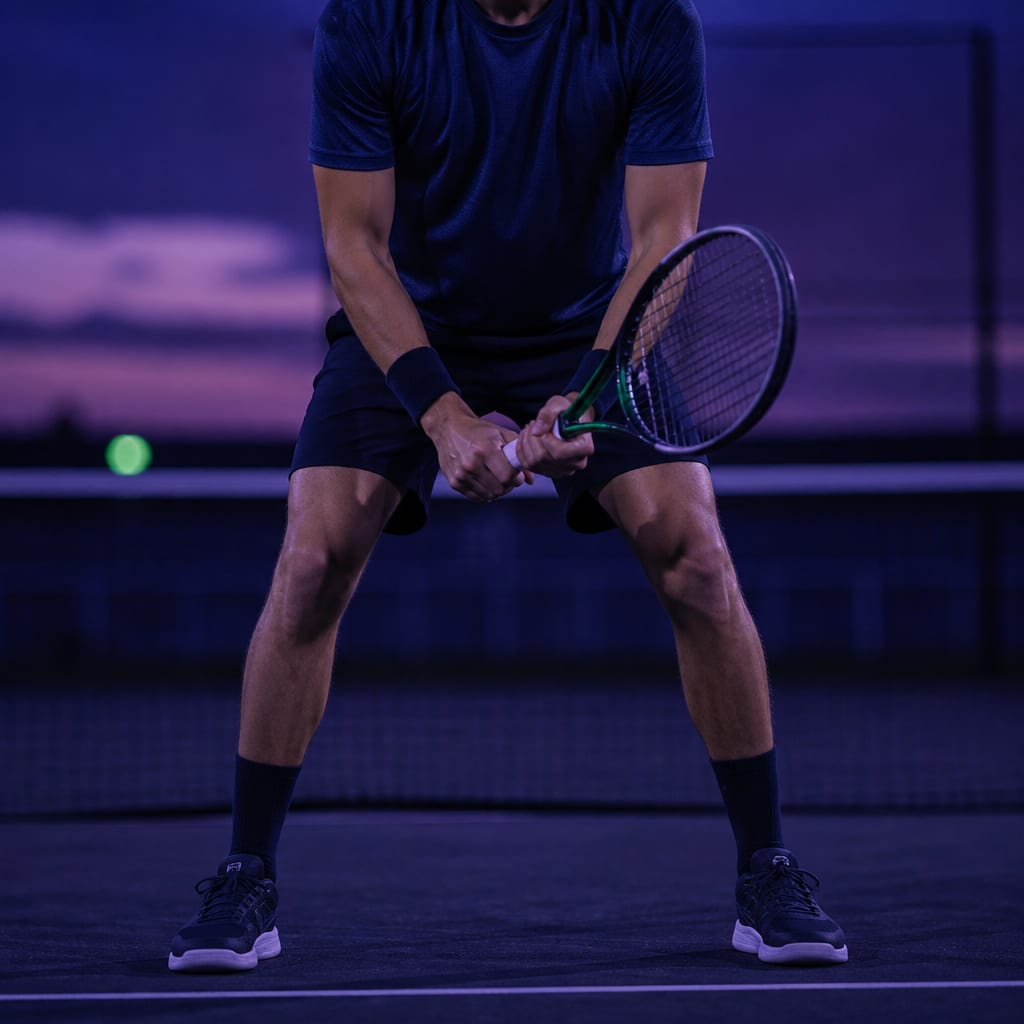 A tennis player in the ready position, feet wider than shoulders, knees flexed, weight forward, racquet held in front with the non-dominant hand on the throat